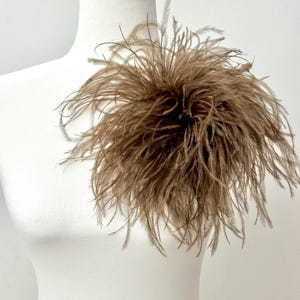 May include: A large, circular decorative accessory made of light brown ostrich feathers. The feathers are arranged in a dense, voluminous cluster, radiating outward from a central point, creating a soft, textured, and layered appearance.