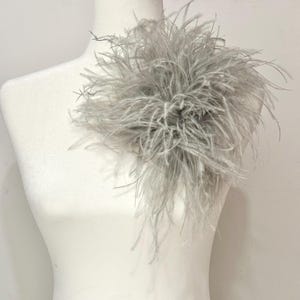 May include: A decorative accessory featuring a cluster of light gray ostrich feathers arranged in a circular, voluminous shape. The soft, wispy plumes extend outward from a central point, creating a textured, layered appearance on a white mannequin.