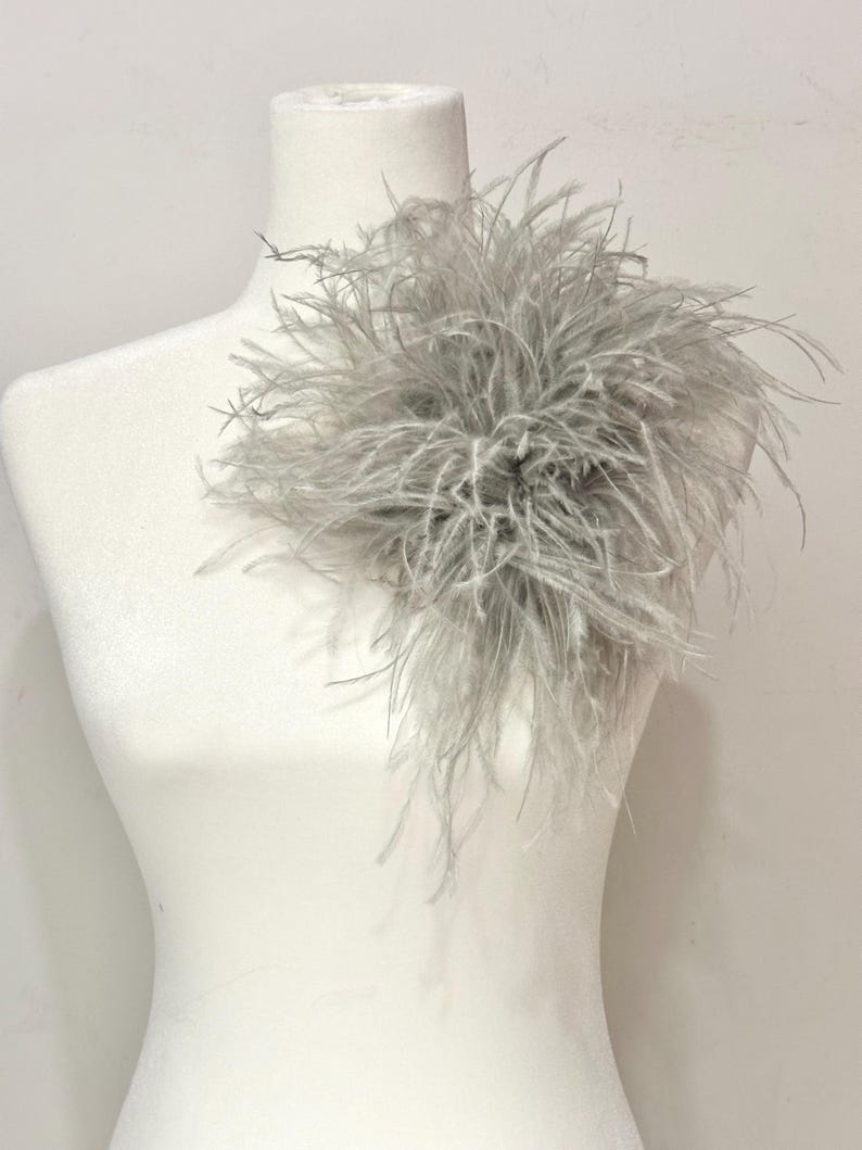 May include: A decorative accessory featuring a cluster of light gray ostrich feathers arranged in a circular, voluminous shape. The soft, wispy plumes extend outward from a central point, creating a textured, layered appearance on a white mannequin.