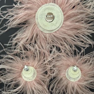 May include: Three pale pink ostrich feather accessories with circular felt backings. The largest piece features a silver-toned pin closure, while the two smaller pieces are equipped with silver-toned metal alligator clips for secure attachment.
