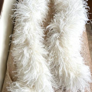 May include: Two fluffy, white feather boas are draped across a surface. The boas are made of soft, delicate feathers, creating a voluminous and textured appearance. The color is a bright, clean white, and the overall effect is elegant and luxurious.