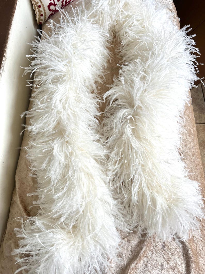 May include: Two fluffy, white feather boas are draped across a surface. The boas are made of soft, delicate feathers, creating a voluminous and textured appearance. The color is a bright, clean white, and the overall effect is elegant and luxurious.