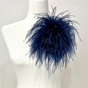 May include: A dark navy blue ostrich feather brooch featuring long, wispy, and delicate plumes arranged in a dense, circular cluster. The feathers radiate outward from a central point, creating a voluminous and textured appearance on the display.