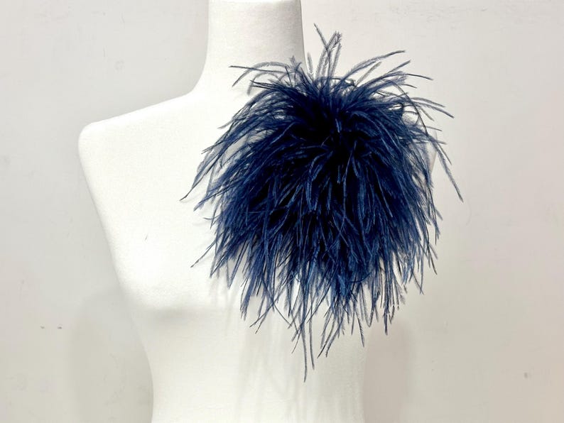 May include: A dark navy blue ostrich feather brooch featuring long, wispy, and delicate plumes arranged in a dense, circular cluster. The feathers radiate outward from a central point, creating a voluminous and textured appearance on the display.