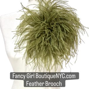 May include: An olive green feather brooch featuring long, wispy plumes arranged in a circular, voluminous cluster. A dark rectangular label is overlaid at the bottom with the white text Fancy Girl BoutiqueNYC.com and Feather Brooch.