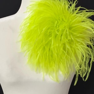 May include: A vibrant, neon green feather accent piece, possibly a shoulder or neckline embellishment, is displayed on a white mannequin. The feathers are densely packed, creating a fluffy, textured appearance against a black background.