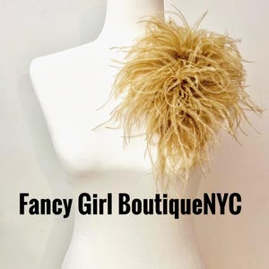 May include: A beige feather accessory attached to a white mannequin torso. The feathers are long, wispy, and clustered together in a dense, rounded shape. The text Fancy Girl BoutiqueNYC is printed in black letters across the lower section.