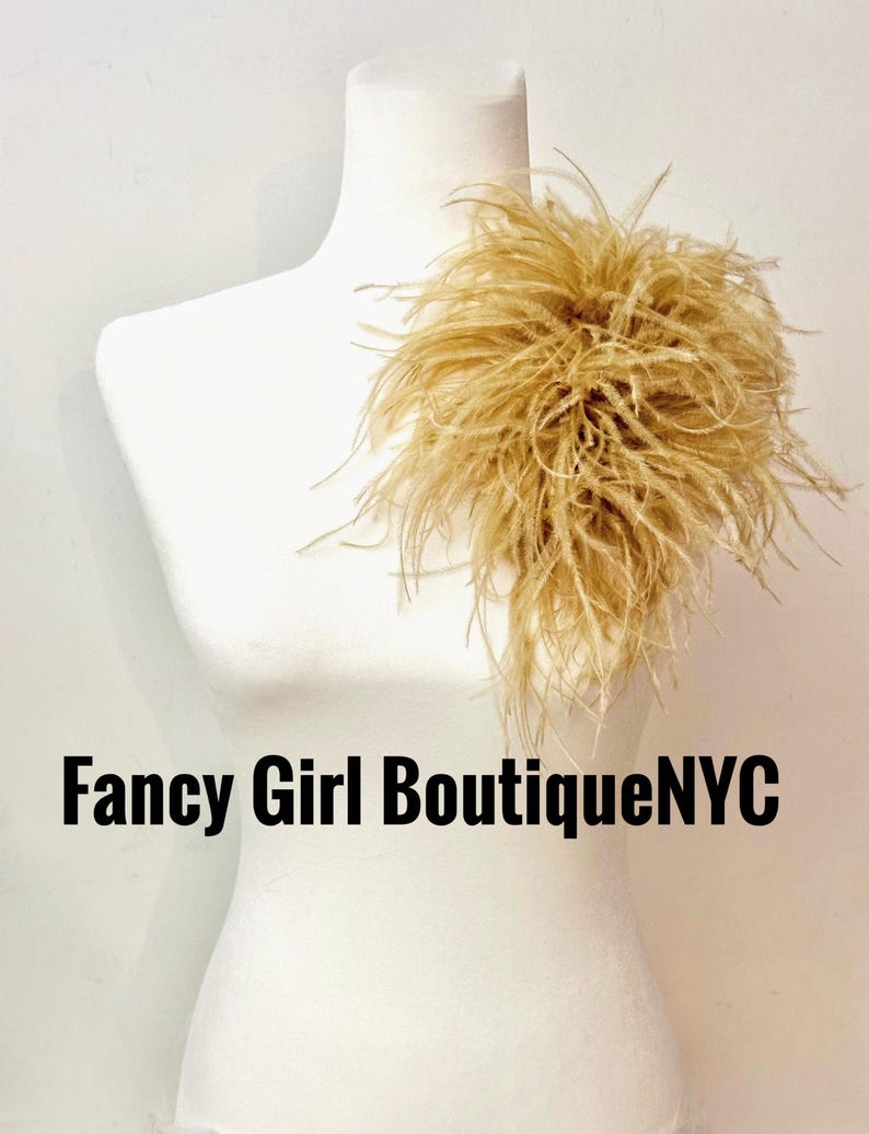 May include: A beige feather accessory attached to a white mannequin torso. The feathers are long, wispy, and clustered together in a dense, rounded shape. The text Fancy Girl BoutiqueNYC is printed in black letters across the lower section.