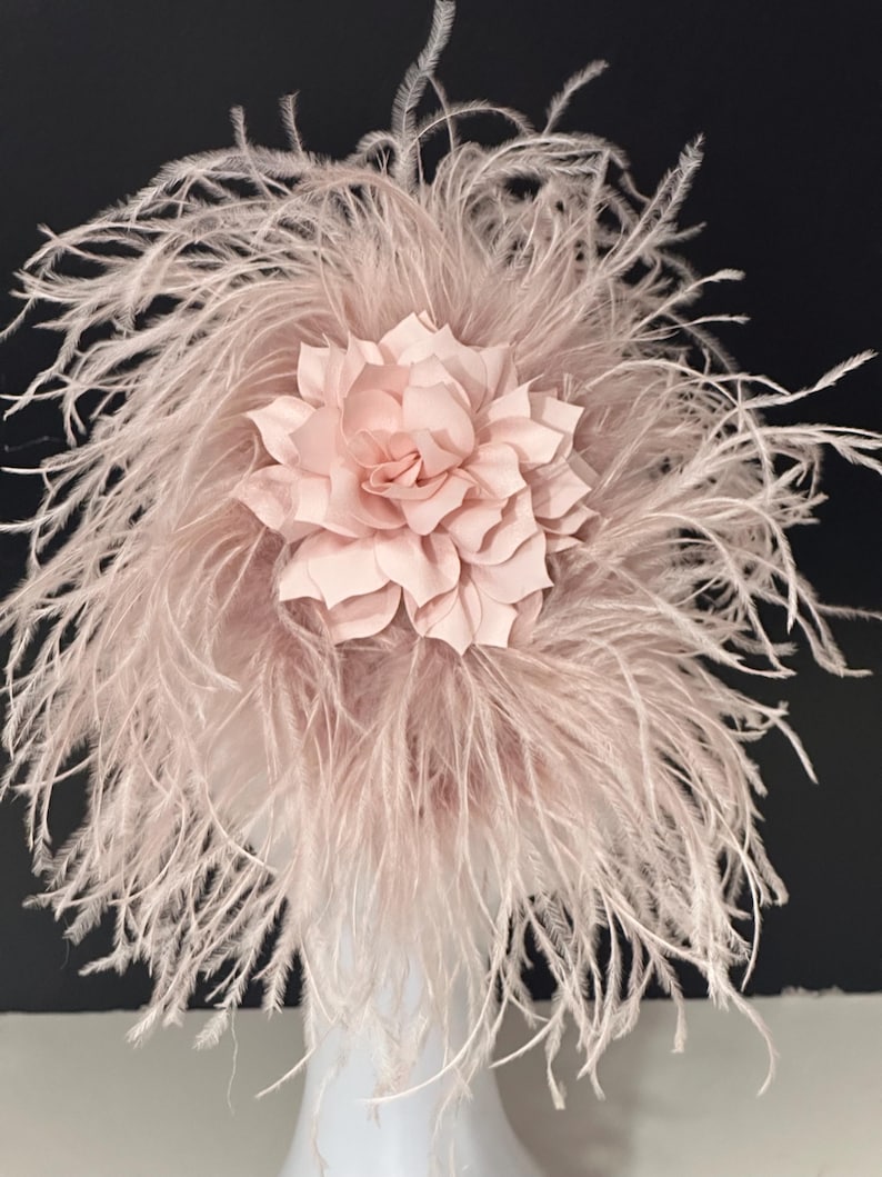May include: A blush pink feather headpiece with a large, layered, blush pink flower.