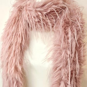 May include: A fluffy, light pink faux fur scarf. The scarf is draped over a white form, showcasing its texture and color. The long, soft fibers create a luxurious and elegant look, perfect for adding a touch of glamour to any outfit.