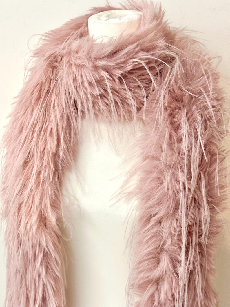 May include: A fluffy, light pink faux fur scarf. The scarf is draped over a white form, showcasing its texture and color. The long, soft fibers create a luxurious and elegant look, perfect for adding a touch of glamour to any outfit.