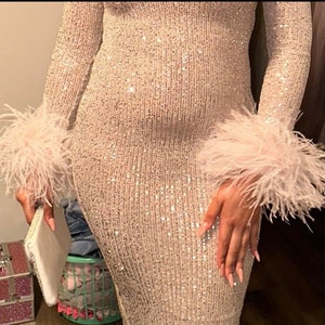 May include: A long-sleeved dress covered in shimmering silver sequins, featuring prominent light pink feather trim at the cuffs. The garment is form-fitting and reaches below the knees, showcasing a dense, reflective texture throughout the fabric.