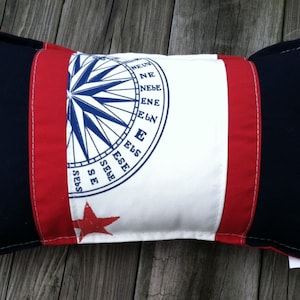 May include: A white pillow with a red and blue nautical compass design. The compass has the directions N, NE, E, SE, S, SW, W, and NW. The pillow has a red star on the bottom left corner.