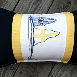 May include: A decorative pillow with a white background and yellow and navy blue trim. The pillow features a stylized illustration of a sailboat with a yellow and blue flag.