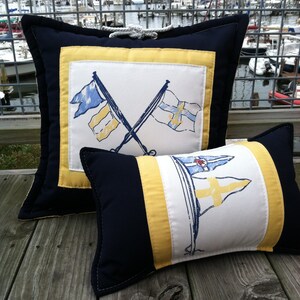 May include: Two decorative pillows with nautical themed designs. The larger pillow is navy blue with a yellow border and features two crossed flags with a white background. The smaller pillow is navy blue with yellow stripes and features a white background with a sailboat and two flags.