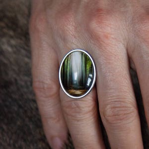 May include: A silver ring with an oval-shaped stone featuring a painted scene of a forest path.