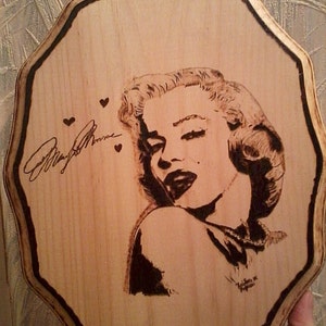 May include: Wooden plaque featuring a pyrography portrait of Marilyn Monroe, with her signature and three small hearts. The artwork is framed by a dark, scalloped edge, and the wood has a light, natural finish.