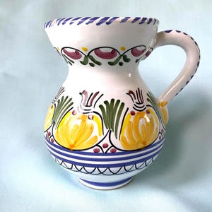 May include: A white ceramic pitcher featuring hand-painted yellow floral motifs with green leaves, accented by purple and blue decorative bands. The piece includes a curved handle and a wide, flared rim with blue painted detailing along the edge.