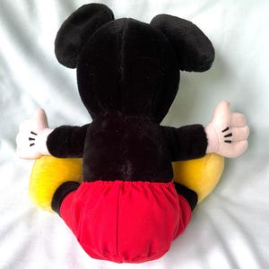 Puede incluir: Un peluche de Mickey Mouse, visto desde atr&aacute;s, con orejas y cuerpo negros, pantalones cortos rojos, zapatos amarillos y manos enguantadas blancas con el pulgar hacia arriba. El juguete es suave y tierno.