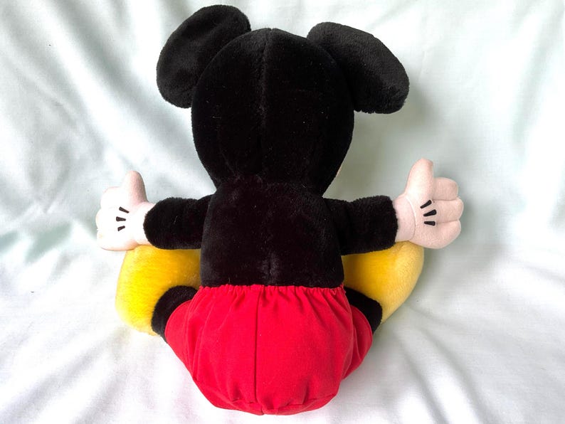 Puede incluir: Un peluche de Mickey Mouse, visto desde atr&aacute;s, con orejas y cuerpo negros, pantalones cortos rojos, zapatos amarillos y manos enguantadas blancas con el pulgar hacia arriba. El juguete es suave y tierno.