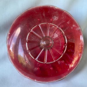 May include: A clear, round, red glass serving dish with a decorative, ridged center and a scalloped edge. The dish has a glossy finish and is likely vintage.