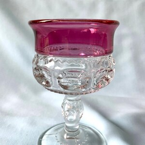 K&ouml;nnte beinhalten: Ein klares Glas mit rubinrotem Rand. Die Schale des Glases weist ein detailliertes, gemustertes Design auf. Der Stiel ist transparent mit einem dekorativen Design, und der Fu&szlig; ist rund.