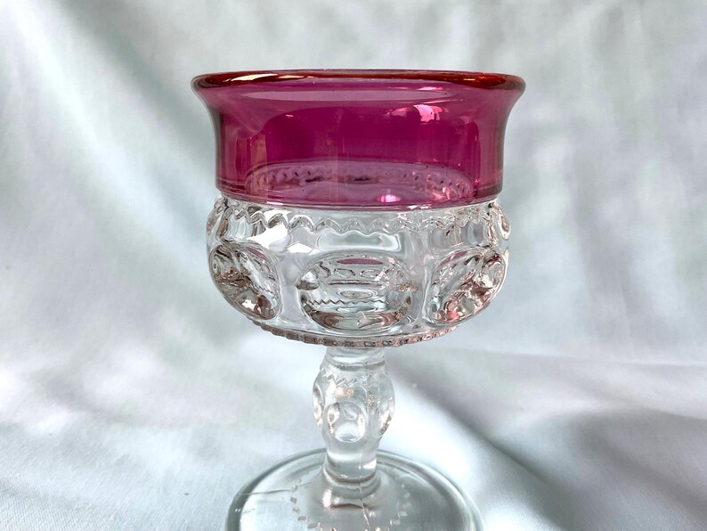 K&ouml;nnte beinhalten: Ein klares Glas mit rubinrotem Rand. Die Schale des Glases weist ein detailliertes, gemustertes Design auf. Der Stiel ist transparent mit einem dekorativen Design, und der Fu&szlig; ist rund.