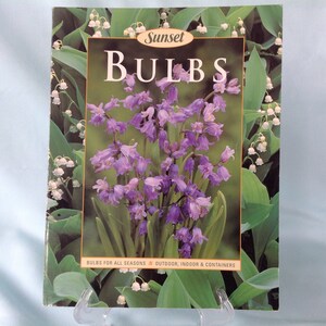 May include: A book cover titled "BULBS" by Sunset, featuring a vibrant image of purple bell-shaped flowers. The cover is framed by green leaves and white flowers, with text at the bottom indicating the book's focus on outdoor, indoor, and container gardening.