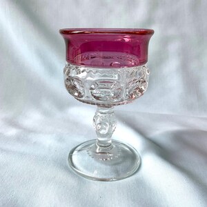 K&ouml;nnte beinhalten: Ein klares Glas mit rubinrotem Rand. Die Schale hat ein detailliertes, gemustertes Design, und der Stiel und der Fu&szlig; sind ebenfalls aus klarem Glas. Das Glas ist wahrscheinlich Vintage und eignet sich zum Servieren von Wein oder anderen Getr&auml;nken.
