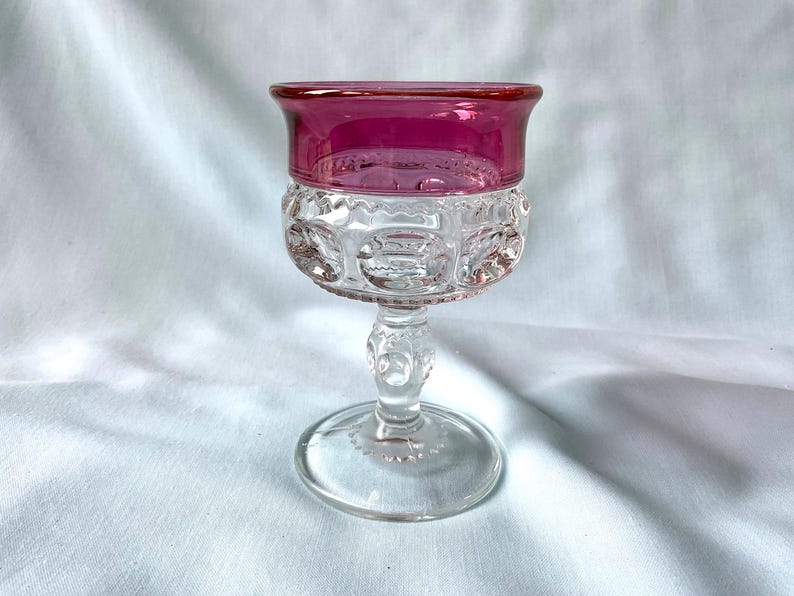 K&ouml;nnte beinhalten: Ein klares Glas mit rubinrotem Rand. Die Schale hat ein detailliertes, gemustertes Design, und der Stiel und der Fu&szlig; sind ebenfalls aus klarem Glas. Das Glas ist wahrscheinlich Vintage und eignet sich zum Servieren von Wein oder anderen Getr&auml;nken.
