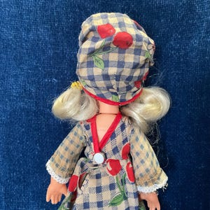 May include: A vintage doll wearing a checkered bonnet with red cherries and a matching dress. The dress has a cherry print and red trim. The doll has blonde hair and is set against a blue background.