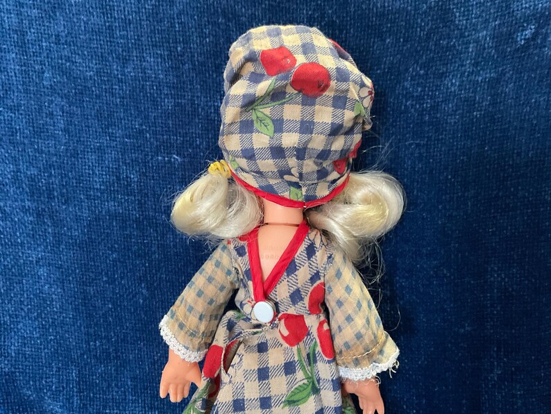 May include: A vintage doll wearing a checkered bonnet with red cherries and a matching dress. The dress has a cherry print and red trim. The doll has blonde hair and is set against a blue background.