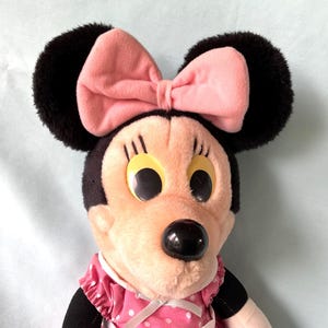 May include: A plush Minnie Mouse doll featuring a pink bow, black ears, and a pink and white polka dot dress. The doll has large, round eyes and a black nose. The overall design is a classic representation of the iconic character.