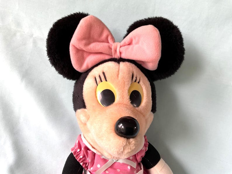 May include: A plush Minnie Mouse doll featuring a pink bow, black ears, and a pink and white polka dot dress. The doll has large, round eyes and a black nose. The overall design is a classic representation of the iconic character.