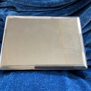May include: A rectangular, silver-colored metal case with a shiny, reflective surface. The word "Radisson" is subtly imprinted on the surface. The case has a clean, minimalist design and is set against a blue background.