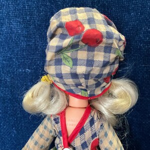 May include: A vintage doll with blonde hair and a checkered bonnet adorned with red cherries. The doll wears a patterned dress with red trim. The overall aesthetic is reminiscent of a classic, retro style.