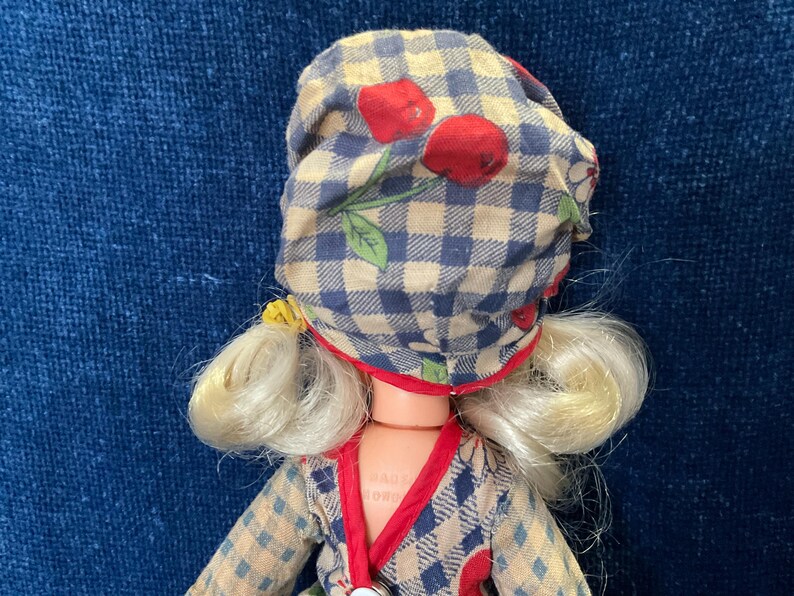 May include: A vintage doll with blonde hair and a checkered bonnet adorned with red cherries. The doll wears a patterned dress with red trim. The overall aesthetic is reminiscent of a classic, retro style.