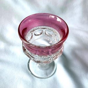 K&ouml;nnte beinhalten: Ein klares Glas mit einem breiten, kreisf&ouml;rmigen Rand in einem tiefrosa Farbton. Die Schale des Kelches weist ein gemustertes Design auf und steht auf einem klaren Stiel. Das Glas steht vor einem wei&szlig;en Hintergrund.
