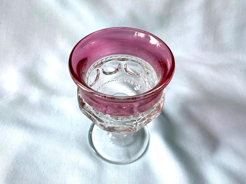 K&ouml;nnte beinhalten: Ein klares Glas mit einem breiten, kreisf&ouml;rmigen Rand in einem tiefrosa Farbton. Die Schale des Kelches weist ein gemustertes Design auf und steht auf einem klaren Stiel. Das Glas steht vor einem wei&szlig;en Hintergrund.