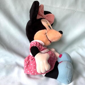 May include: A plush Minnie Mouse doll, featuring black fur, a pink bow, and a pink polka-dot dress. The doll has a smiling expression and light blue shoes. The doll is sitting on a light blue surface.