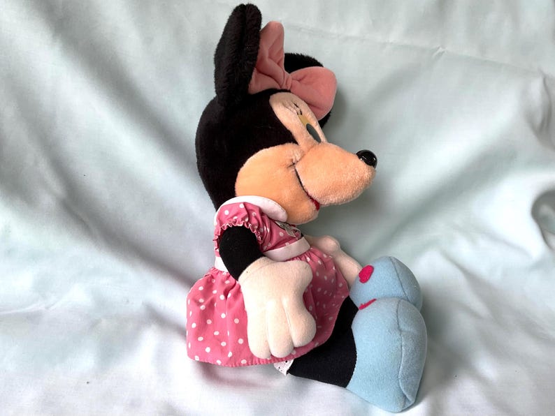 May include: A plush Minnie Mouse doll, featuring black fur, a pink bow, and a pink polka-dot dress. The doll has a smiling expression and light blue shoes. The doll is sitting on a light blue surface.
