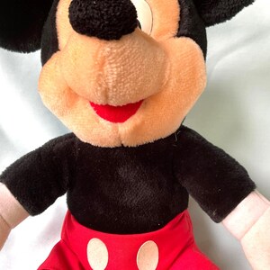 Puede incluir: Un peluche de Mickey Mouse. El juguete tiene un cuerpo negro, orejas y una nariz redonda. La cara es de color canela con una boca roja. Mickey lleva pantalones cortos rojos con dos c&iacute;rculos blancos.