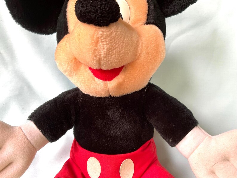 Puede incluir: Un peluche de Mickey Mouse. El juguete tiene un cuerpo negro, orejas y una nariz redonda. La cara es de color canela con una boca roja. Mickey lleva pantalones cortos rojos con dos c&iacute;rculos blancos.