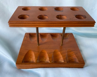 May include: A vintage wooden pipe rack with eight circular openings on top and six curved slots below. The rack is supported by metal rods and has a warm, brown finish, showcasing the natural wood grain.