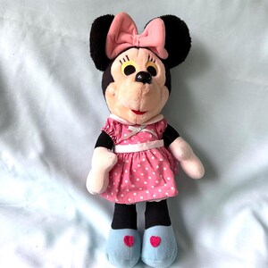 May include: A Minnie Mouse plush doll with black ears, a pink bow, and a pink polka-dot dress. The doll has black legs, white hands, and blue shoes with red heart accents. The doll is approximately 12 inches tall.