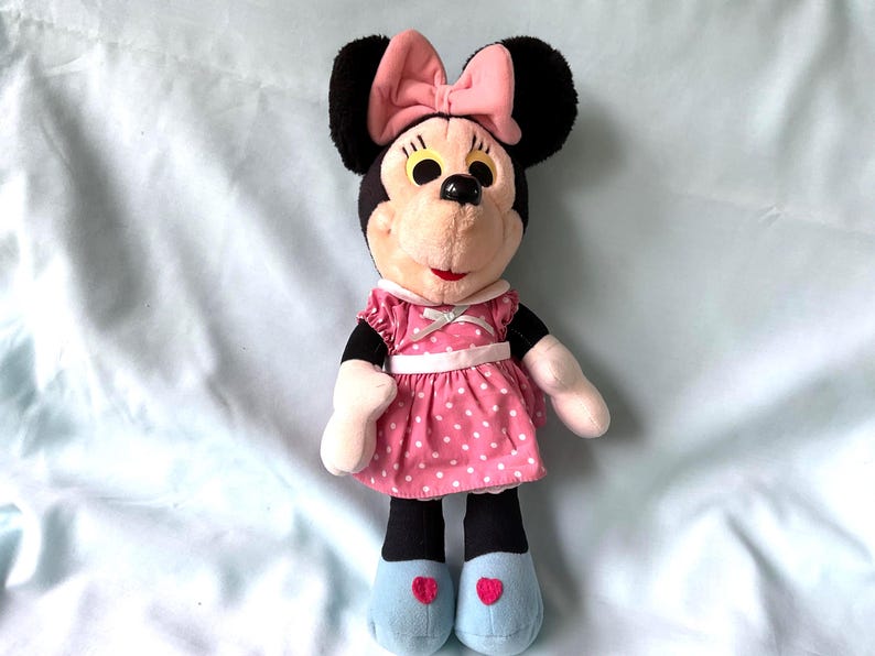 May include: A Minnie Mouse plush doll with black ears, a pink bow, and a pink polka-dot dress. The doll has black legs, white hands, and blue shoes with red heart accents. The doll is approximately 12 inches tall.