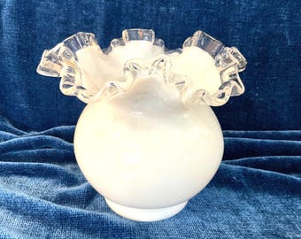 May include: A white milk glass vase with a spherical body and a wide, ruffled, clear glass rim. The vase features a smooth, opaque finish on the main body, contrasting with the translucent, wavy edge that creates a decorative, scalloped opening.