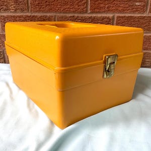 May include: A bright orange plastic storage case with a square shape and a textured top surface. The container features a single gold-toned metal latch on the front center for closure and an integrated handle built into the top lid design.