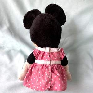 May include: A plush Minnie Mouse doll, viewed from the back, features a black head with large ears and a pink dress with white polka dots. The dress has a white collar and a white belt.