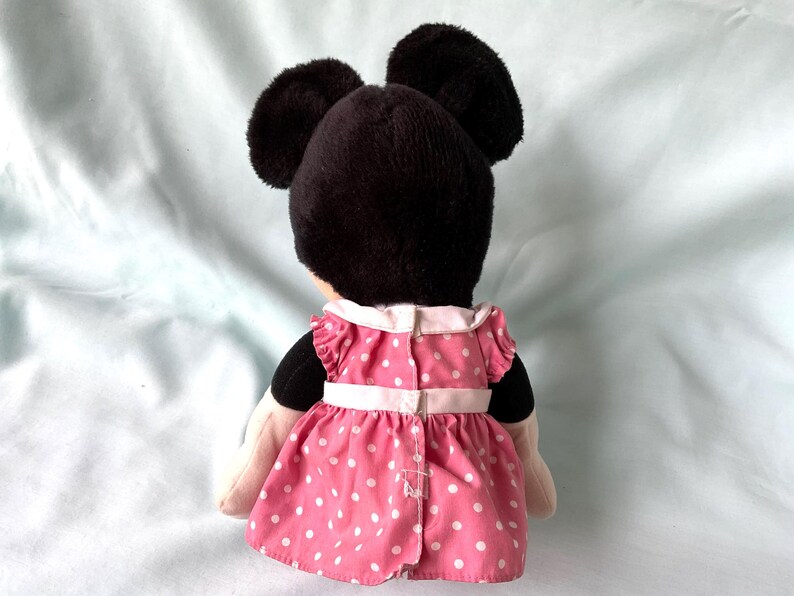 May include: A plush Minnie Mouse doll, viewed from the back, features a black head with large ears and a pink dress with white polka dots. The dress has a white collar and a white belt.
